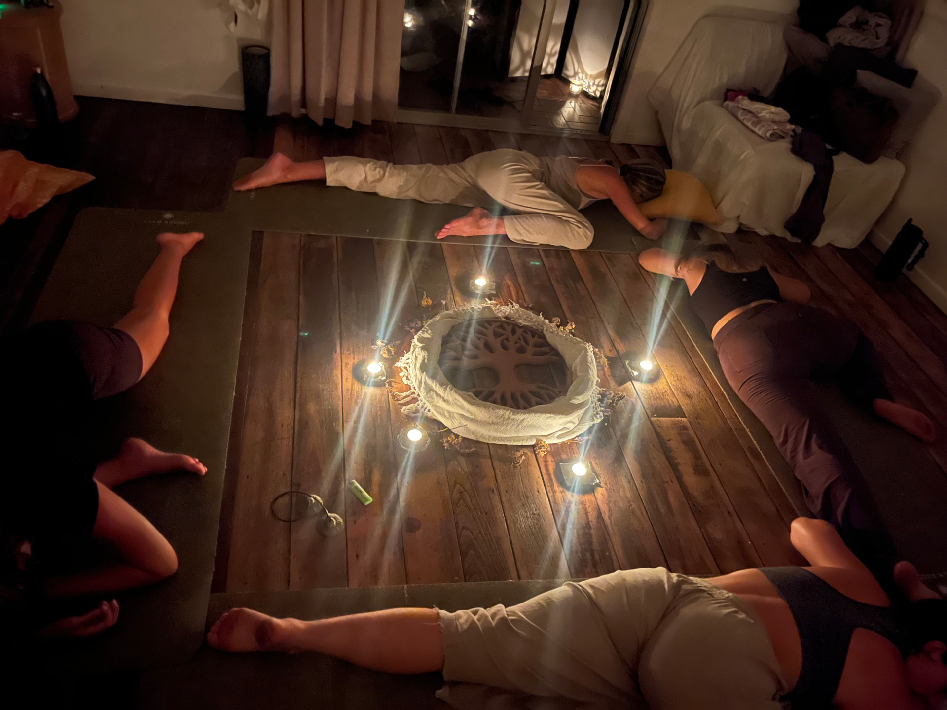 Women resting in a candlelit circle around a woven mandala on a wooden floor