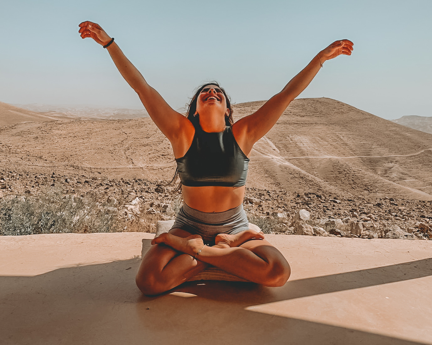 Michal Magen seated in lotus pose with arms open to the sky in a sunlit desert landscape